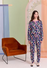 Tumedas sinise põhivärviga lillemustriga pidžaama, millel on pikad varrukad ja püksid ning roosad detailid ja nööriga pingutus. Kõrval on oranž kangast tugitool.