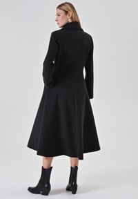 Schwarzer Mantel mit ausgestelltem Design, langen Ärmeln und weitem Kragen. Trägt man über schwarzen Knöchelstiefeln, vor hellem Hintergrund präsentiert.