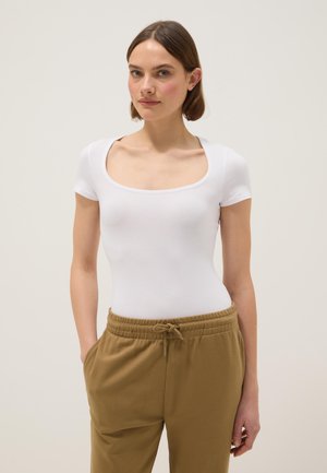 Vrouw met kort bruin haar, gekleed in een wit aansluitend T-shirt met ronde hals en beige broek met trekkoord, staand met handen in de zakken.