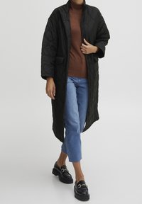 Abrigo largo negro acolchado con cuello mandarín y dos bolsillos delanteros, usado sobre un suéter de cuello alto marrón y vaqueros azules cortos, combinado con zapatos negros.