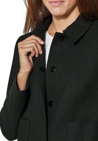 Losjes vallende donkergroene jas van gestructureerde stof, met grote zwarte knopen en zijzakken. Heeft een klassieke kraag en een zachte voering.