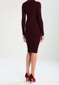 Robe moulante bordeaux côtelée avec manches longues, ourlet à hauteur du genou et silhouette ajustée ; portée avec des chaussures rouges à talons hauts.