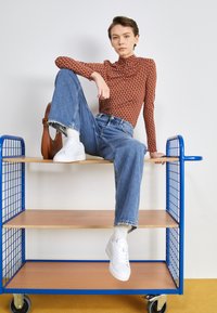 Maglietta a maniche lunghe marrone con pattern, jeans blu a gamba larga e sneakers bianche. Carrello in legno con telaio blu in metallo e ruote girevoli.