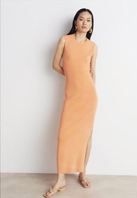 Oransje ermeløs maxikjole med en sidesliss. Glatt, elastisk stoff. Modellen har på seg beige flip-flops og hoopsøreringer. Enkel, tettsittende design.
