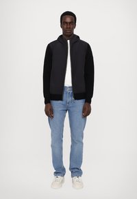 Jachetă neagră cu fermoar și glugă, cu manșete tricotate, purtată peste o cămașă albă. Blugi din denim de culoare albastru deschis și adidași albi completează ținuta.