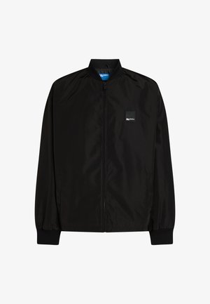 Melna bomber jakna, izgatavota no gludas, vieglas auduma. Izceļas ar priekšējo rāvējslēdzēju, ribotiem piedurknēm un nelielu logo uzšuvumu uz krūšu.