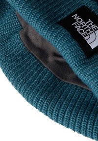 Strikket hue i teal med en ribbet tekstur. Har en grå, blød foring og et sort patch med hvid branding på siden.