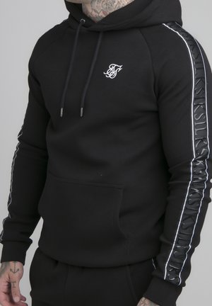 Homme portant un sweat à capuche noir avec un logo blanc sur la poitrine et un texte marqué le long des manches, poche kangourou avant visible.