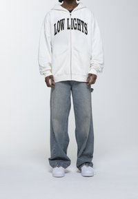Sudadera blanca con cremallera y texto "LOW LIGHTS" en negro en el pecho, combinada con unos vaqueros de pierna ancha azul claro y zapatillas blancas.