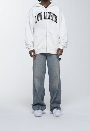 Sweat à capuche zippé blanc avec le texte "LOW LIGHTS" en noir sur la poitrine, associé à un jean large bleu clair et des baskets blanches.
