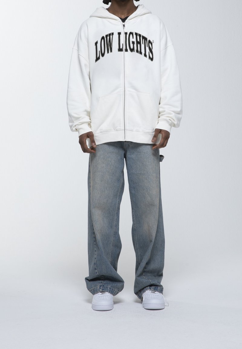 Sudadera blanca con cremallera y texto "LOW LIGHTS" en negro en el pecho, combinada con unos vaqueros de pierna ancha azul claro y zapatillas blancas.