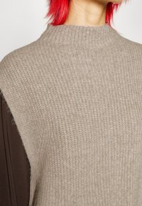 Maglione lavorato a maglia beige con collo alto, caratterizzato da un motivo a coste testurizzato. Il capo ha una vestibilità rilassata con maniche marroni che emergono.