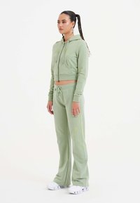 Lichtgroene, korte zip-up hoodie met een voorzak, gecombineerd met bijpassende broek met een zijfloral patroon. Stevige witte sneakers.