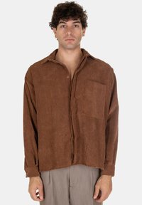 Camicia in velluto a coste marrone con bottoni frontali, una singola tasca sul petto e maniche lunghe, caratterizzata da una texture a coste e una vestibilità rilassata.