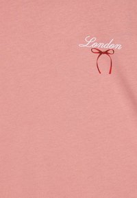 Camiseta rosa claro con la palabra "Londres" en un estilo de escritura blanca y un pequeño gráfico de un lazo rojo. Textura suave con un diseño casual.