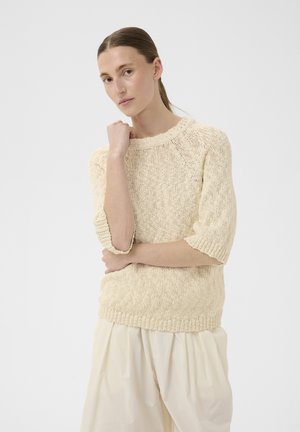 Donna con i capelli pettinati all'indietro che indossa un maglione in maglia color crema con maniche al gomito e pantaloni plissettati larghi di colore bianco sporco, in piedi su uno sfondo semplice.
