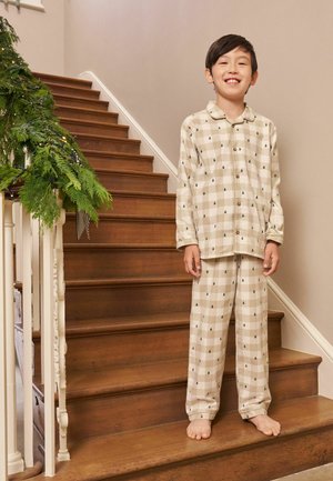 Beige ternet børnepyjamas sæt med sorte træmønstre, knap-op top og bukser med elastisk talje. Trætrappe i baggrunden.