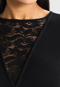 Pull noir avec un design en V, doté d'une superposition en dentelle noire détaillée au niveau du col et d'un tissu doux et texturé dans l'ensemble.