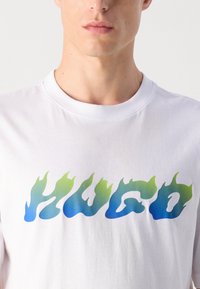 Lähikuva henkilöstä, joka pitää yllään valkoista pyöreäkaulaista t-paitaa, jonka rinnassa on sinivihreä gradienttityylinen "HUGO"-teksti.