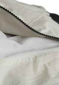 Tejido blanco con cremallera y forro de malla texturizada. Presenta herrajes de cremallera negros y un exterior arrugado. Diseño limpio y minimalista.