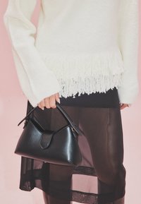 Črna usnjena torbica s strukturirano obliko in kratkim ročajem, ki jo drži oseba v belem puloverju s frendami in črni prosojni krilu.