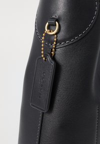 Primer plano de la esquina de un bolso de cuero negro con anilla dorada y etiqueta negra de cuero Coach sujeta con cadena de cuentas dorada.