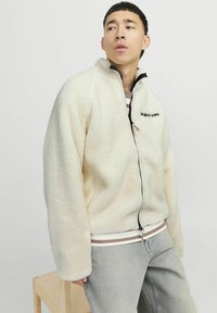 Kreemist fleece-jakk, millel on lukuga esiots, must logo rinnal ja kontrastsete triibuliste ribadega äär. Mudel on paaritatud heledate teksadega.