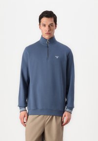 Μπλε φούτερ με φερμουάρ από μαλακό ύφασμα, με ψηλό γιακά, ελαφριές λωρίδες στα μανίκια και ένα μικρό λογότυπο στο στήθος.