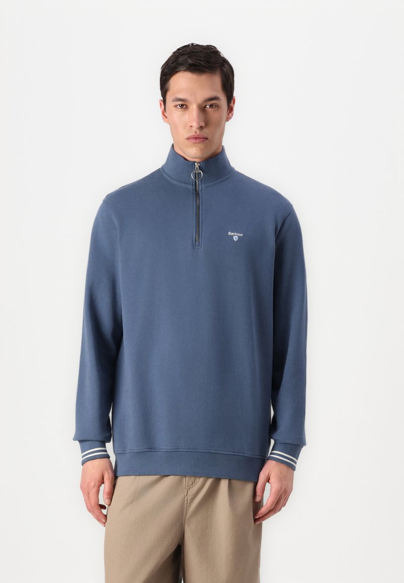 Μπλε φούτερ με φερμουάρ από μαλακό ύφασμα, με ψηλό γιακά, ελαφριές λωρίδες στα μανίκια και ένα μικρό λογότυπο στο στήθος.