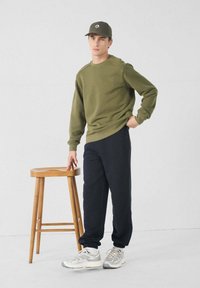 Hombre con sudadera verde oliva, pantalones de chándal negros y zapatillas grises, de pie junto a un taburete de madera y tocándolo, frente a un fondo liso.