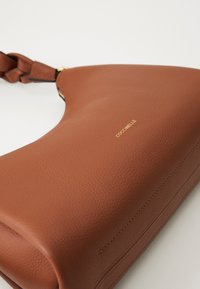 Brown leather handbag with braided strap and gold "COCCINELLE" logo on smooth textured surface against a white background.