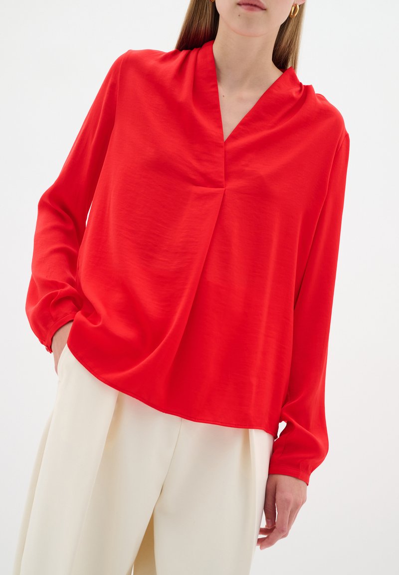 Blusa rossa a maniche lunghe con scollo a V, vestibilità comoda e polsini arricciati. Tessuto liscio con una sottile texture. Abbinata a pantaloni chiari.