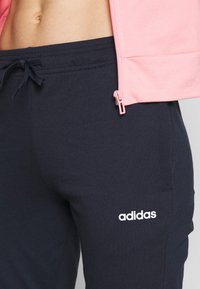 Talia i biodra w czarnych spodniach dresowych Adidas z sznurkiem oraz część różowej kurtki zapinanej na zamek, odsłaniające nagi brzuch.