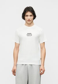 Νέος άνδρας με μεσαίου μήκους σκουρόχρωμα μαλλιά που φοράει λευκό μπλουζάκι με το κείμενο "Replay Custom Service" και γκρι παντελόνι, στέκεται μπροστά σε απλό φόντο.