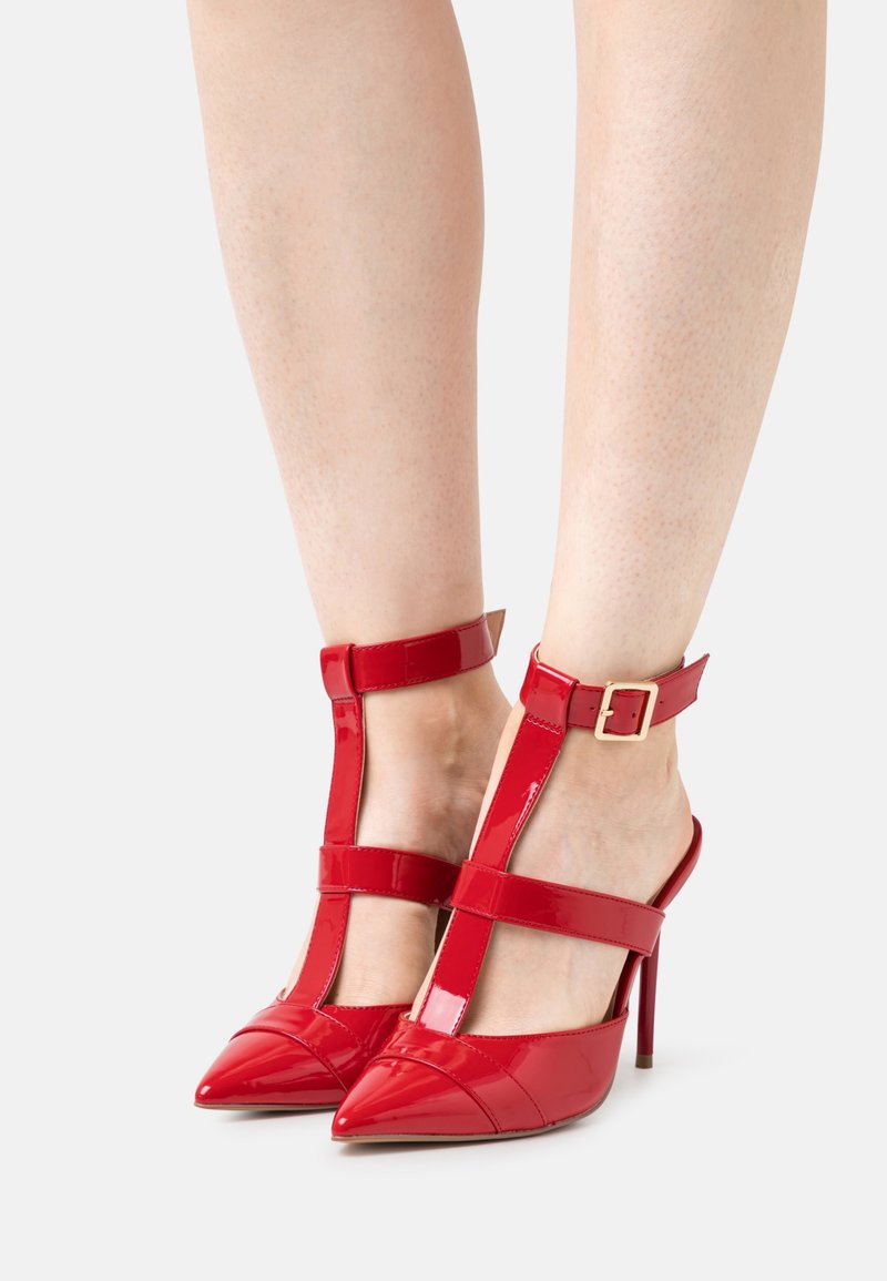 Chaussures à talons hauts en cuir verni rouge brillant avec brides à la cheville et au pied, fermées par des boucles dorées sur des jambes nues.