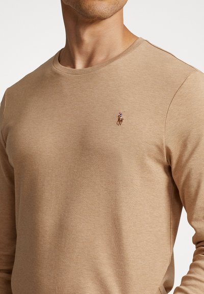 Beige langemouwen shirt van zacht materiaal met een ronde halslijn. Heeft een geborduurd logo op de linkerborst. Strak, lichtgewicht ontwerp.