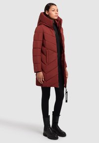 Roter isolierter Steppmantel mit großer Kapuze, diagonalem Chevron-Design, Reißverschluss vorne, Seitentaschen und einem Anhänger am Reißverschluss.