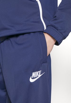 Person iført navyblåt Nike træningssæt med hånd i lommen, der har hvid Nike-logo og lynlåsdetalje på jakken.