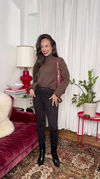 Γυναίκα που χαμογελά φορώντας καφέ ζιβάγκο, μαύρο παντελόνι και μπότες, στέκεται σε σαλόνι με κόκκινο βελούδινο καναπέ, μοτίβο χαλί, κόκκινο φωτιστικό και φυτά σε γλάστρες.