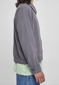 Šedá bavlněná bunda s límcem, bočními kapsami a zapínáním na knoflíky na manžetách, nošená přes světle zelenou košili a modré džíny. Hladká textura.