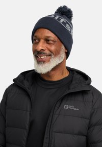 Black puffer jacket with a zip, featuring a logo patch. Dark blue knitted beanie with a pom-pom and light gray text across the brim.