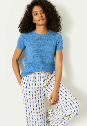 Frau mit lockigem Haar steht mit einer Hand in der Tasche, sie trägt ein blaues, kurzärmeliges Stricktop und weiße Hose mit blauem Blattmuster.