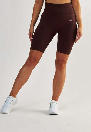 Shorts marron à côtes avec une taille haute, arborant un petit logo en forme de cœur. Portés avec des chaussures de sport blanches sur un fond uni.