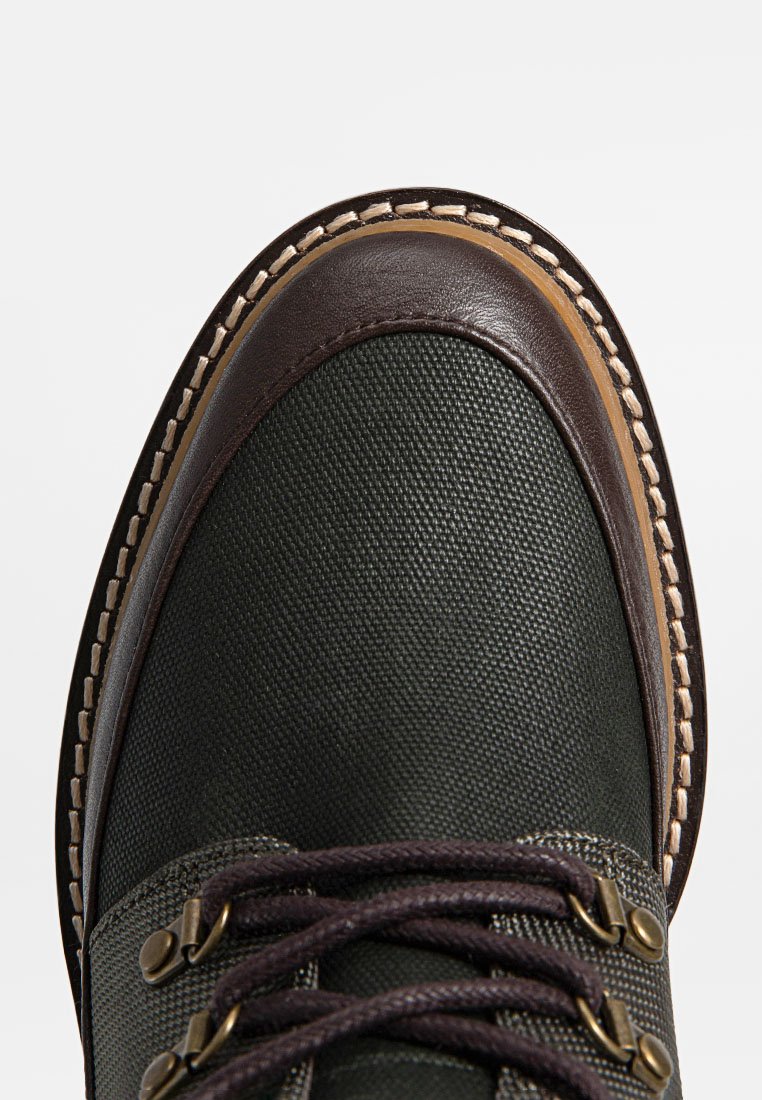 Brązowa skórzana cholewka i ciemnozielona teksturowana tkanina, z okrągłymi brązowymi sznurówkami, metalowymi oczkami i kontrastowym beżowym przeszyciem wzdłuż krawędzi.
