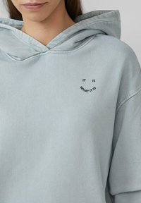Lichtblauwe hoodie met een ontspannen pasvorm, trekkoordkapuchon en gebogen tekst "HET IS WAT HET IS" geborduurd in het zwart op de borst.