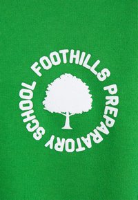 Zelena tkanina s bijelim kružnim logotipom s ikonom drveta i tekstom "Pripremna škola Foothills" oko njega. Glatka tekstura.