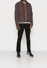 Giacca trapuntata marrone scuro con tasche a zip e colletto rialzato, indossata sopra un felpe marrone e jeans neri, abbinata a sneakers nere.