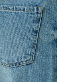 Tela vaquera en azul claro, con un bolsillo trasero izquierdo con costuras beige y una textura ligeramente descolorida.