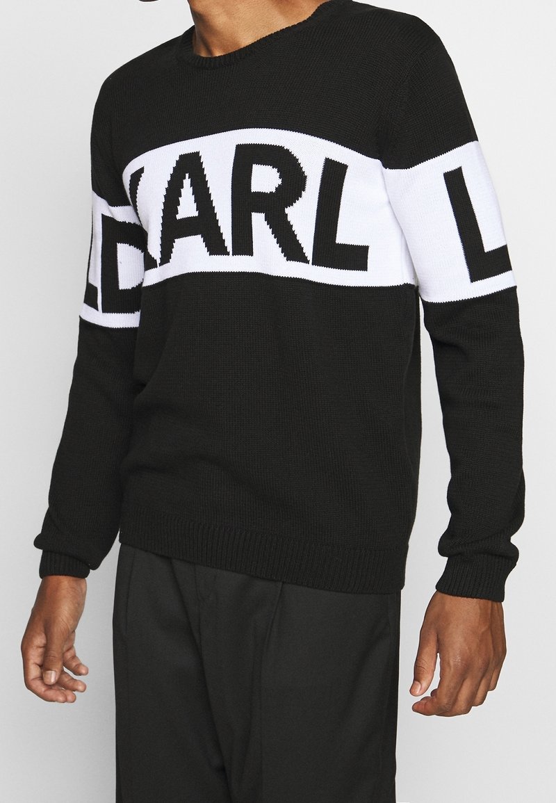 Homme portant un pull noir avec une large bande blanche sur la poitrine et les bras, avec des lettres noires en gras formant « KARL L ».