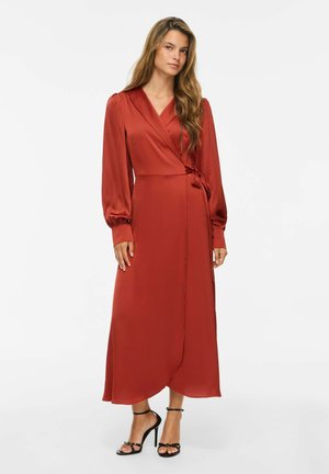 Femme aux longs cheveux portant une robe portefeuille en satin couleur rouille à manches longues et des sandales à talons noirs sur fond blanc.